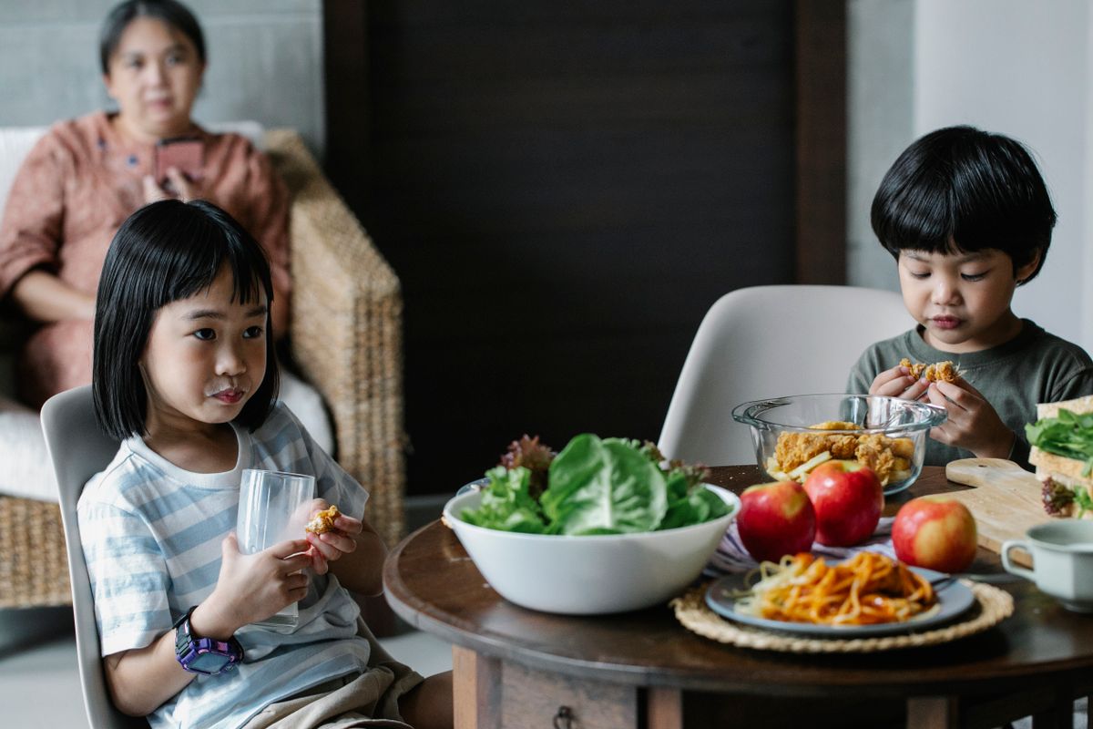 Dos niños desayunando en la cocina con su abuela al fondo